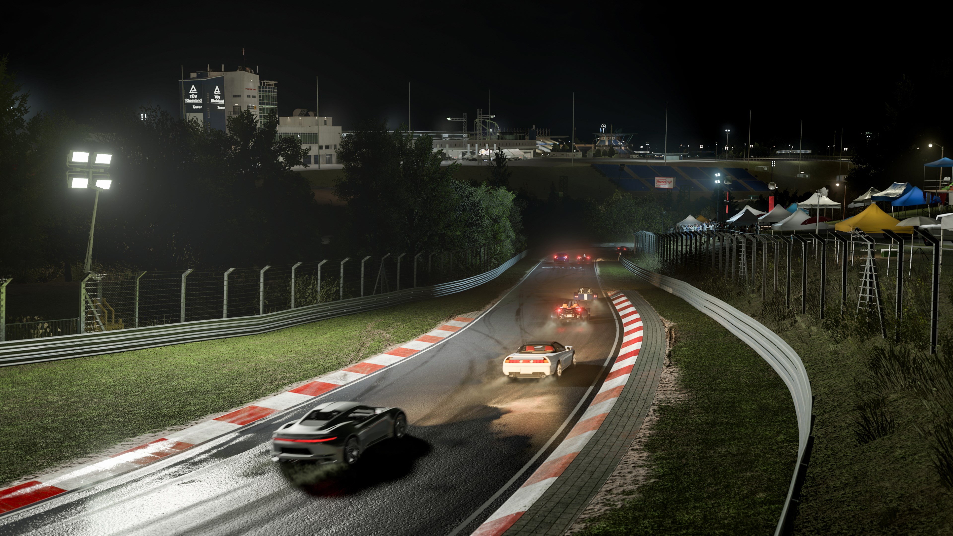 Sports cars racing at night on a wet, winding track with illuminated grandstands and tents in the background.